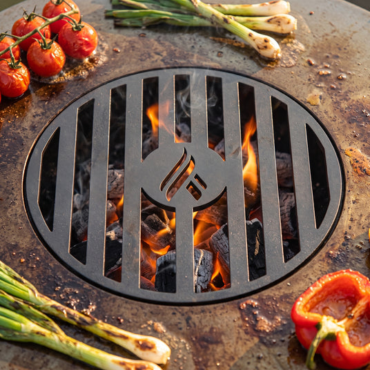 Close-up of the Arteflame laser-cut center grill grate over glowing charcoal, with tomatoes, green onions and pepper on the seasoned griddle