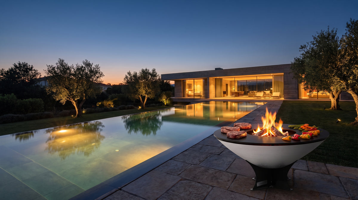 Arteflame 40-inch Platinum Series stainless steel fire pit with cooktop beside an infinity pool at a modern home at dusk