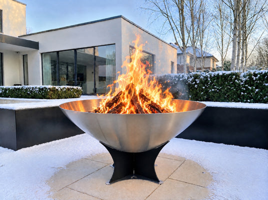 Arteflame 40-inch stainless steel fire pit with a roaring wood fire in a snowy modern garden