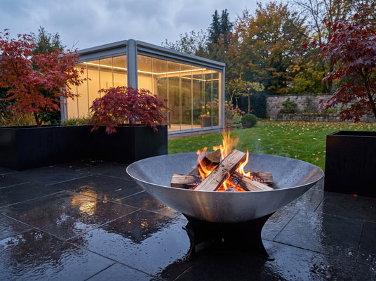 Arteflame 40-inch Platinum Series stainless steel fire pit with raindrops on the bowl, burning on a wet basalt terrace beside a glass garden pavilion