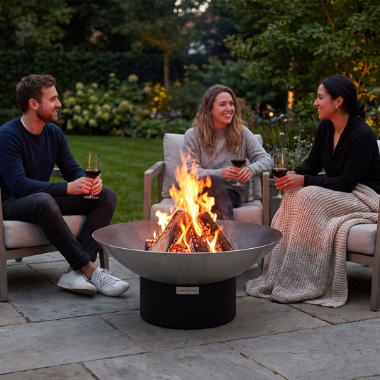 Three friends with wine relaxing around an Arteflame 40-inch Platinum Series stainless steel fire pit on a black low round base on an evening patio