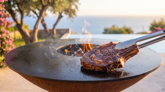 Thick bone-in ribeye searing on the Arteflame grill's steel cooktop beside the wood fire