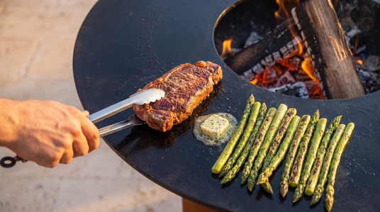 Flipping a seared New York strip steak next to asparagus on the Arteflame cooktop