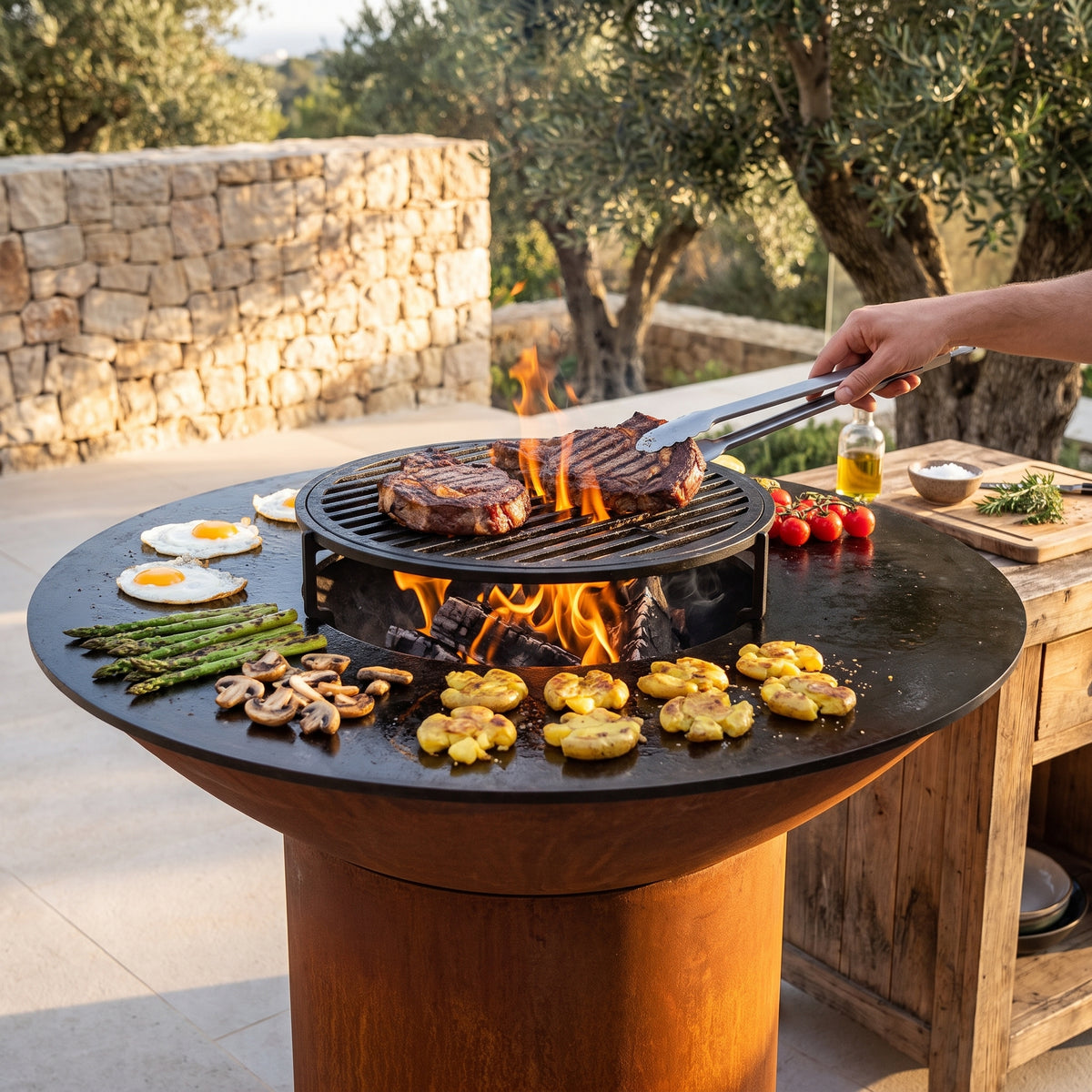 Steaks searing on a grill grate over the wood fire of an Arteflame grill with a round Corten steel base, with eggs, asparagus and potatoes on the flat-top griddle
