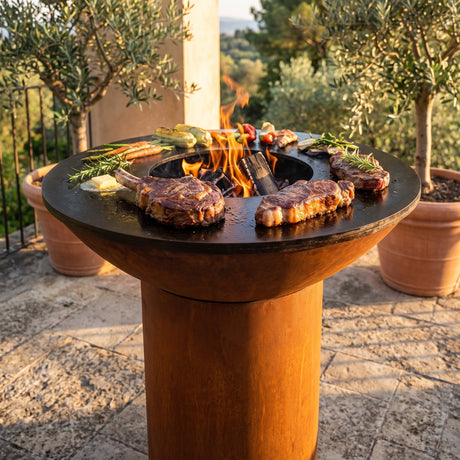 Ribeye and New York strip steaks searing on an Arteflame wood-fired flat-top grill with a round Corten steel base