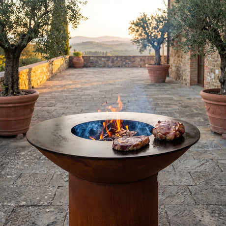 Adding wood-smoke flavor to steak and pork shoulder on an Arteflame wood-fired flat-top grill with a round Corten steel base