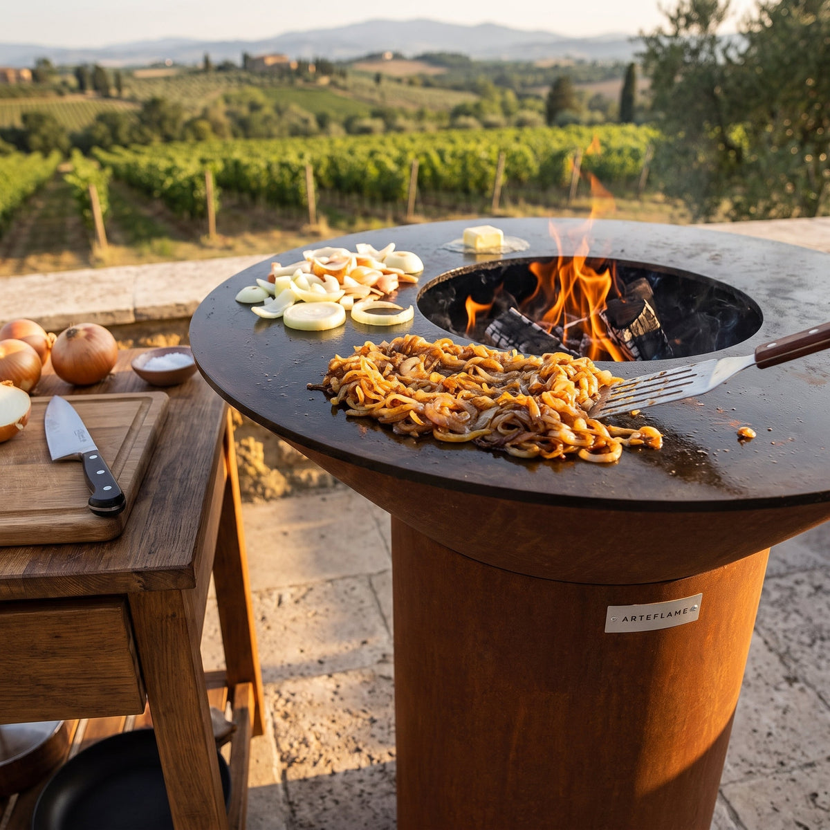 Golden caramelized onions and fresh sliced onions cooking on an Arteflame flat-top grill with a round Corten steel base, overlooking a vineyard
