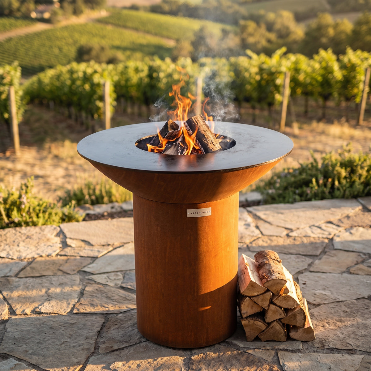 Oak, hickory and cherry firewood stacked beside an Arteflame wood-fired flat-top grill with a round Corten steel base
