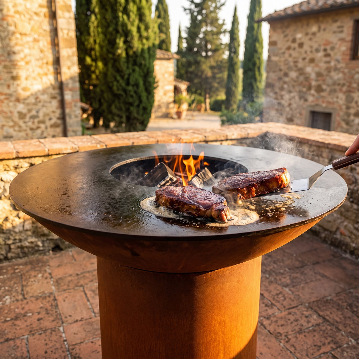 Strip steaks developing a steakhouse crust on an Arteflame wood-fired flat-top grill with a round Corten steel base