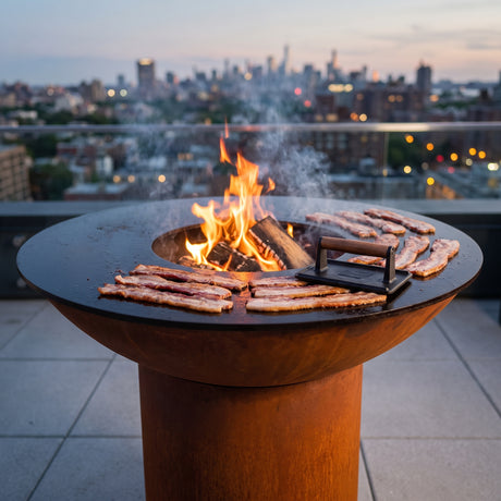 Thick-cut bacon crisping under a bacon press on an Arteflame wood-fired flat-top grill with a round Corten steel base