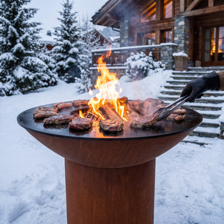 Grilling steaks and sausages in the snow on an Arteflame wood-fired flat-top grill with a round Corten steel base