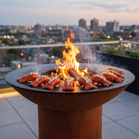 Lobster tails grilling in garlic butter on an Arteflame wood-fired flat-top grill with a round Corten steel base