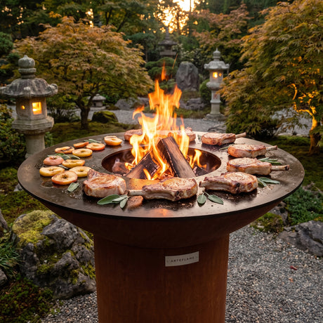 Thick bone-in pork chops searing on an Arteflame wood-fired flat-top grill with a round Corten steel base