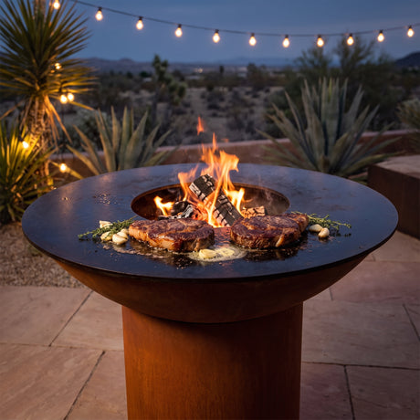 Thick ribeye steaks searing in garlic thyme butter on an Arteflame wood-fired flat-top grill with a round Corten steel base