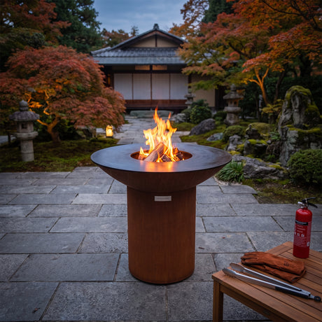 Fire extinguisher, gloves and tongs set out beside an Arteflame wood-fired flat-top grill with a round Corten steel base