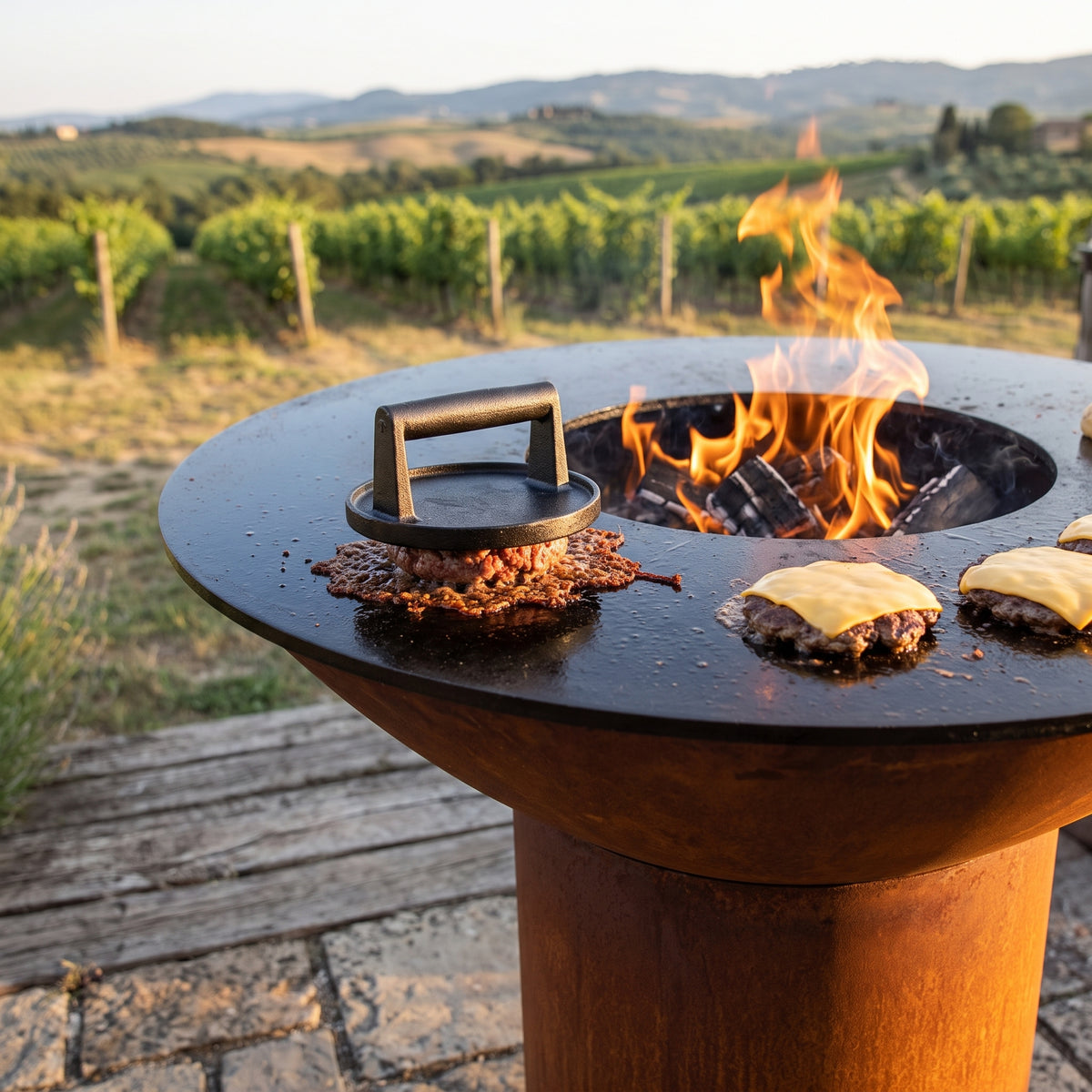 Smash burgers pressed flat with crispy edges on an Arteflame wood-fired flat-top grill with a round Corten steel base
