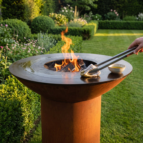 Oiling the hot cooktop with a cloth held in tongs on an Arteflame wood-fired flat-top grill with a round Corten steel base