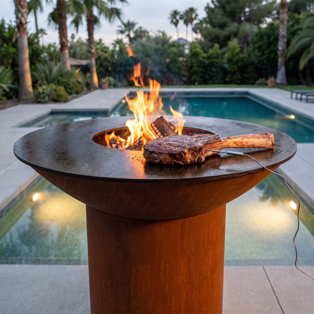Thick tomahawk steak finishing a reverse sear on an Arteflame wood-fired flat-top grill with a round Corten steel base