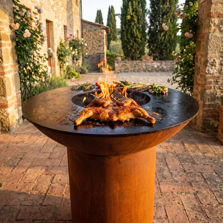 Spatchcocked chicken grilling skin-side down on an Arteflame wood-fired flat-top grill with a round Corten steel base