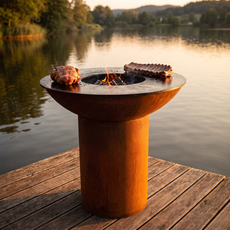 Low and slow pork shoulder and ribs over gentle embers on an Arteflame wood-fired flat-top grill with a round Corten steel base
