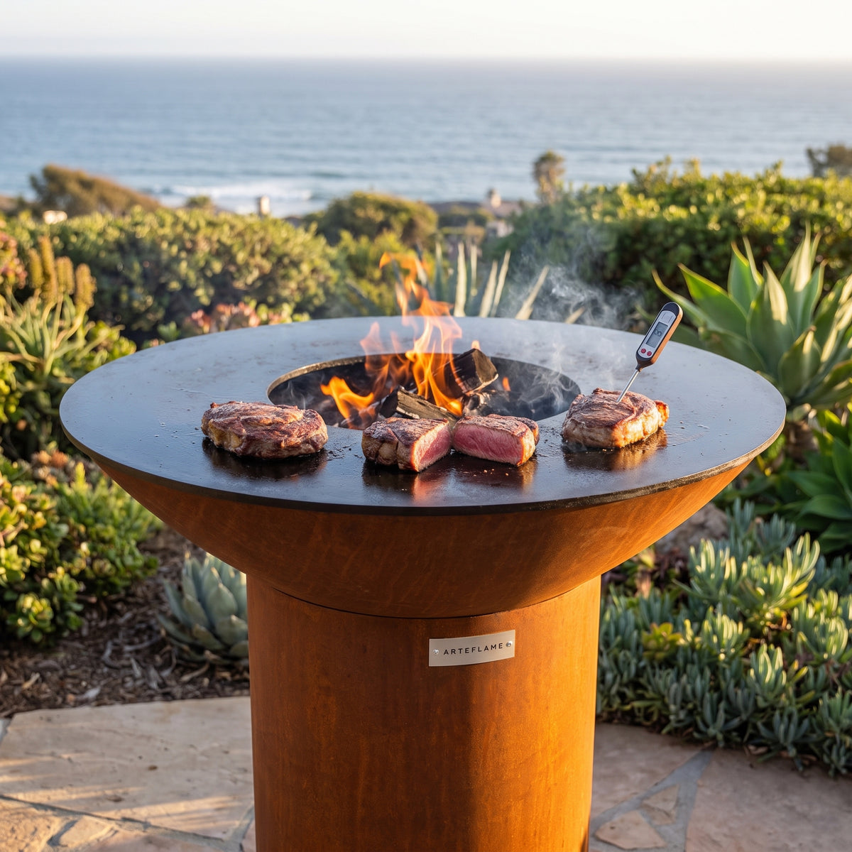 Steaks grilled to medium-rare with a thermometer on an Arteflame wood-fired flat-top grill with a round Corten steel base
