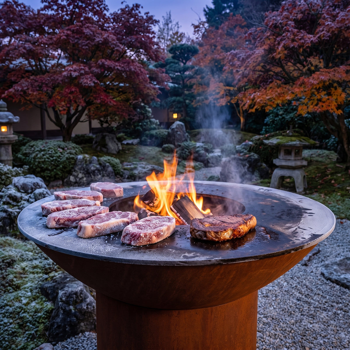 Frozen steaks searing on an Arteflame wood-fired flat-top grill with a round Corten steel base