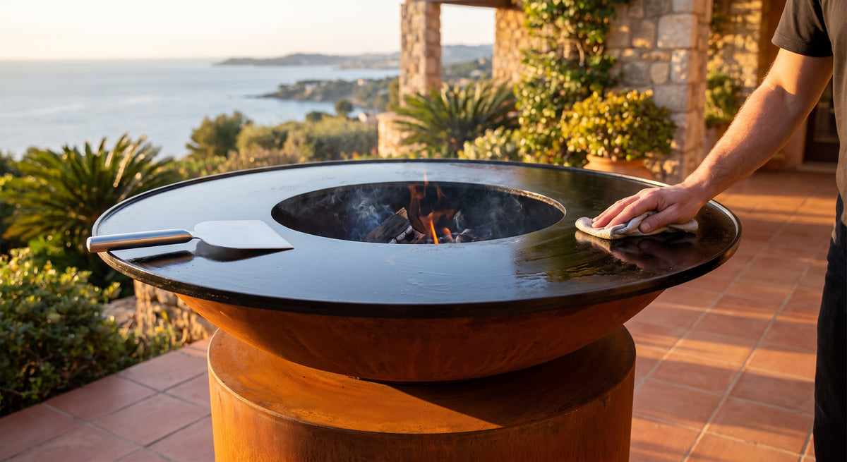 Scraping and wiping a seasoned cooktop without a wire brush on an Arteflame wood-fired flat-top grill with a round Corten steel base