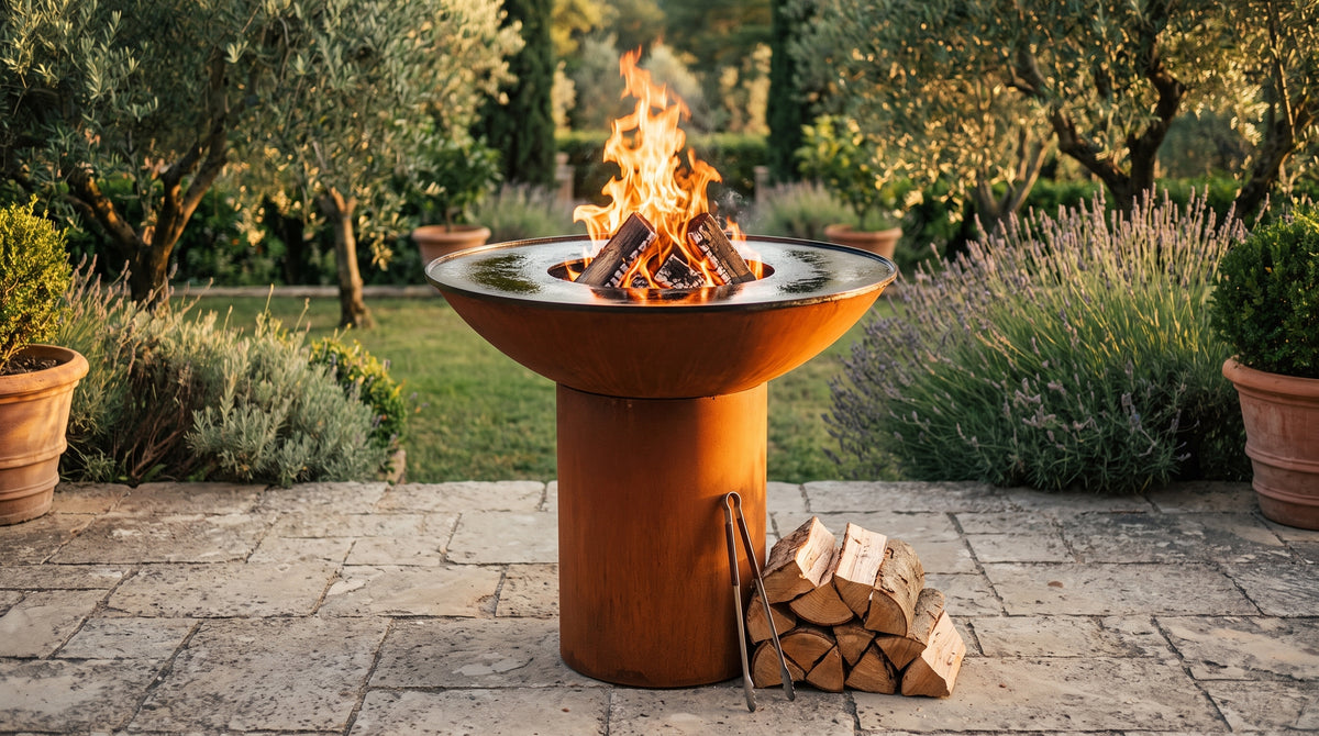Arteflame grill with a round Corten steel base and curved fire bowl, split oak logs burning in the center opening of the empty steel cooktop, with stacked firewood and tongs beside it in a Mediterranean garden