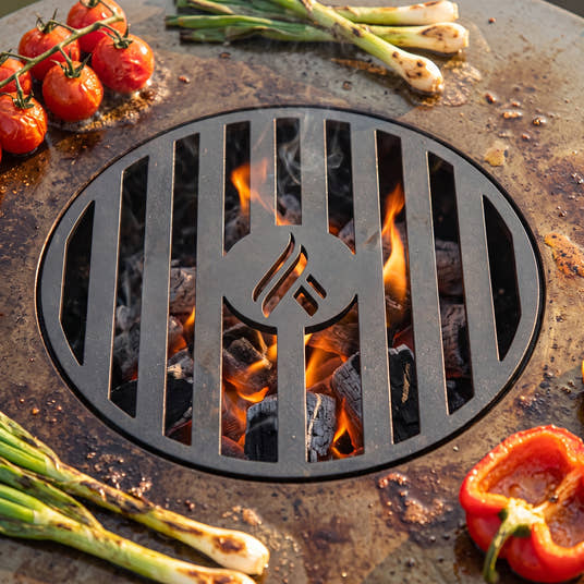 Close-up of the Arteflame laser-cut center grill grate over glowing charcoal, with tomatoes, green onions and pepper on the seasoned griddle