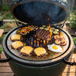 Arteflame Two-Piece Flat-Top Kit on a green kamado grill with smash burgers, shrimp, vegetables and a fried egg on the griddle ring and a ribeye searing on the center grate