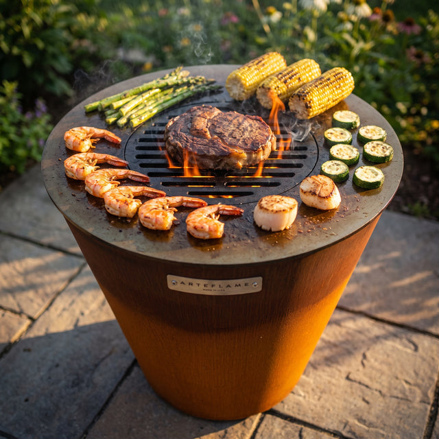 Arteflame ONE20 corten steel grill with shrimp, scallops, asparagus, corn and zucchini on the flat cooktop and a ribeye searing on the center grate