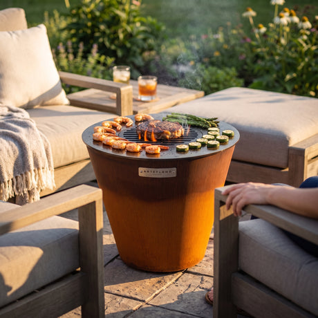Arteflame ONE20 corten steel grill between two cushioned lounge chairs on a garden terrace, cooking shrimp, steak, asparagus and zucchini at armrest height