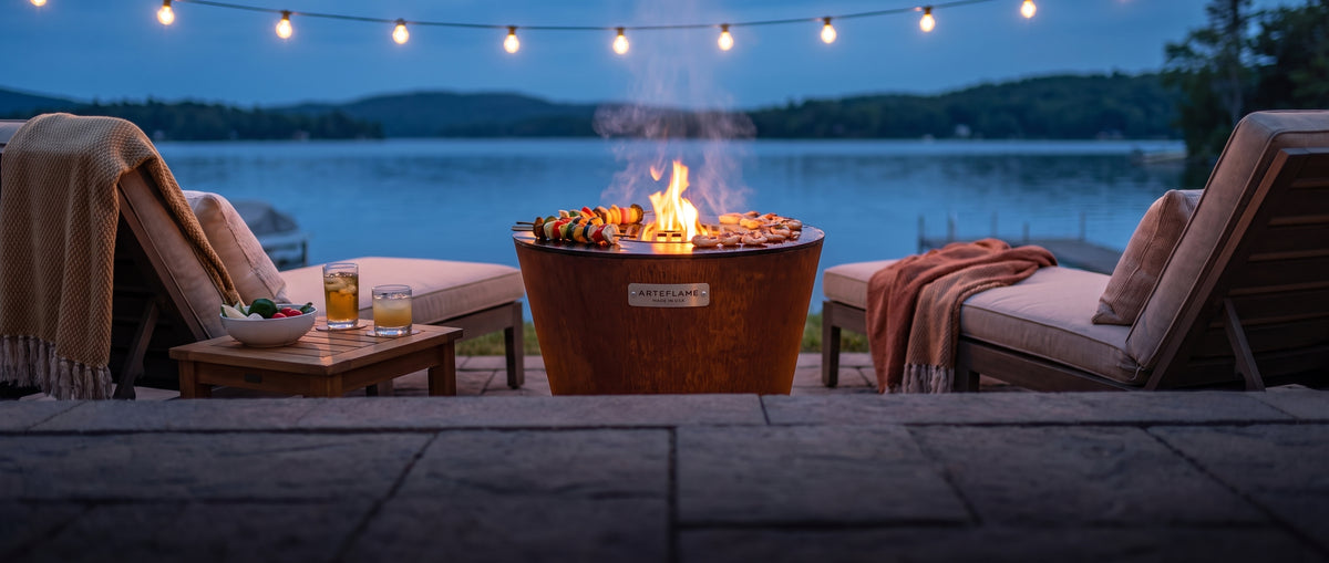 Arteflame ONE20 corten steel grill with skewers and shrimp on the cooktop between two lounge chairs on a lakeside patio at dusk