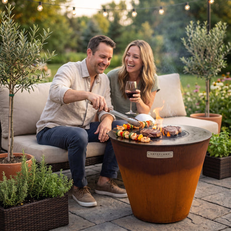Couple seated on an outdoor sofa grilling skewers and steaks on the Arteflame ONE20 right beside them, without getting up
