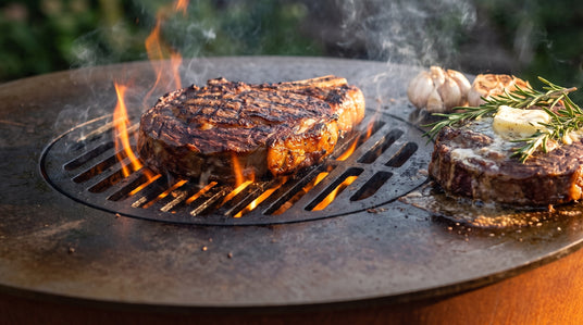 Ribeye searing over open flame on the center grill grate of an Arteflame ONE20 corten steel grill, with a second steak resting on the flat carbon-steel cooktop