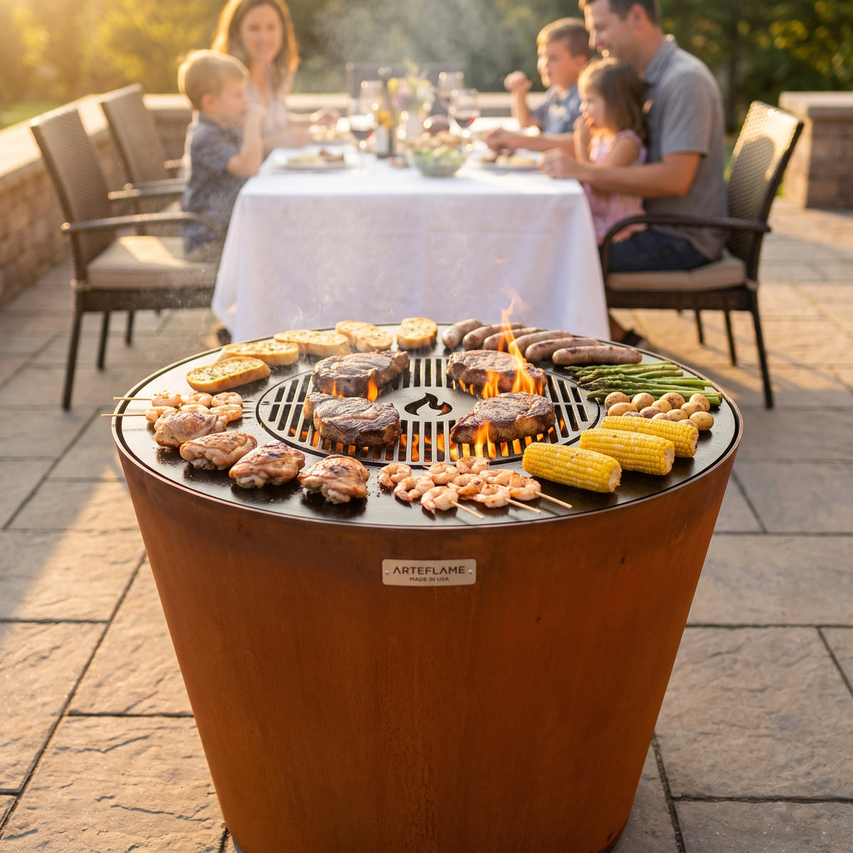 Arteflame ONE30 corten steel grill cooking a family meal at once: steaks on the center grate, chicken, sausages, shrimp, potatoes, corn and garlic bread on the flat cooktop