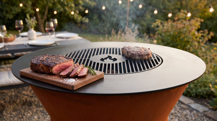 Sliced medium-rare ribeye on a walnut board on the Arteflame ONE30 cooktop, a second steak searing on the center grill grate
