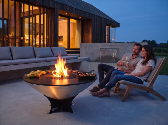 Couple with wine beside the Arteflame 40-inch Platinum Series stainless steel fire pit with cooktop in a contemporary courtyard at blue hour