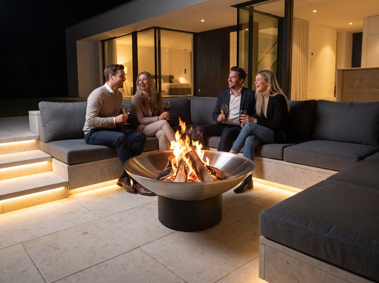 Friends on a modern outdoor sofa around the Arteflame 40-inch Platinum Series stainless steel fire pit, cooktop removed for an open fire at night