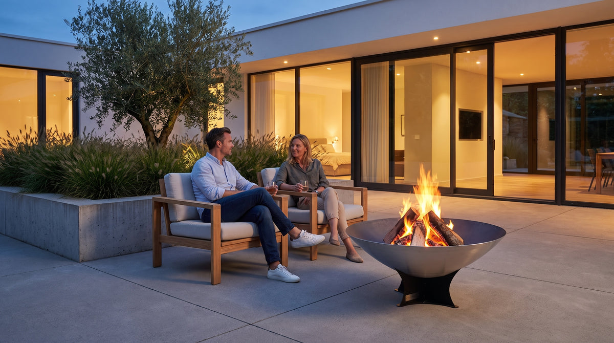 Couple relaxing with drinks beside an Arteflame 40-inch Platinum Series stainless steel fire pit on a black low Euro base in a modern courtyard at dusk