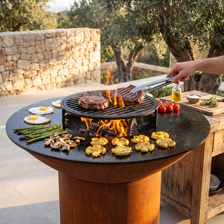 Steaks searing on a grill grate over the wood fire of an Arteflame grill with a round Corten steel base, with eggs, asparagus and potatoes on the flat-top griddle